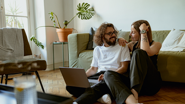 Ein junges Paar sitzt auf dem Fußboden seiner Wohnung mit Laptop auf dem Schoss.