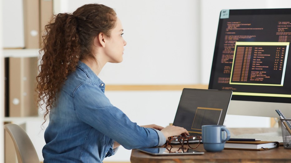 Eine junge Frau gibt am Schreibtisch Daten in den Laptop ein, die sie an einem großen zusätzlichen Bildschirm prüft.