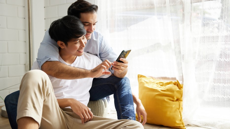 Zwei junge Männer betrachten lächelnd ein Smartphone. Der Arm des eines Mannes liegt auf der Schulter des anderen.