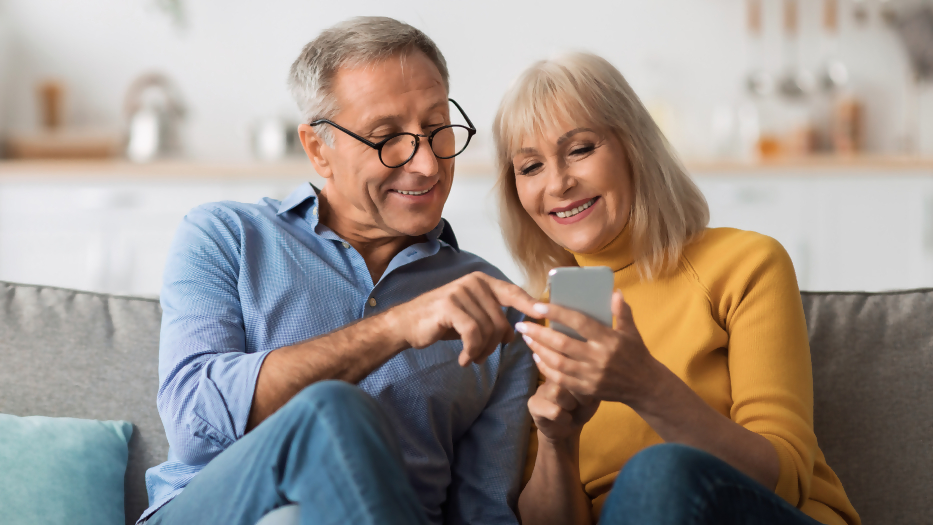 Ein Mann und eine Frau sitzen auf dem Sofa und schauen gemeinsam lächelnd auf ein Smartphone in der Hand der Frau.