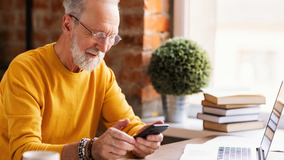Ein Mann mit gelbem Pullover sitzt am Schreibtisch vor dem Laptop und lächelt, während er etwas in sein Smartphone eingibt.