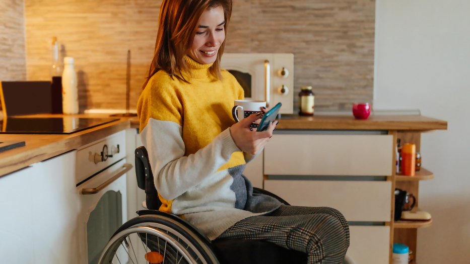 Eine Rollstuhlnutzerin bedient in ihrer Küche mit einer Hand ein Smartphone. In der anderen Hand hält sie eine Tasse.