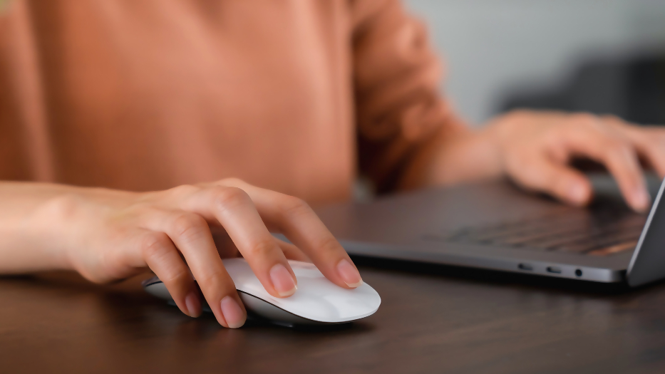 Eine Person arbeitet an einem Schreibtisch mit ihrem Laptop und einer Mouse. Der Bildfokus liegt auf den Händen der Person.